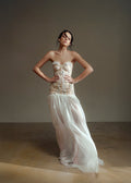 Woman wearing a strapless top and flowing white skirt against a plain background