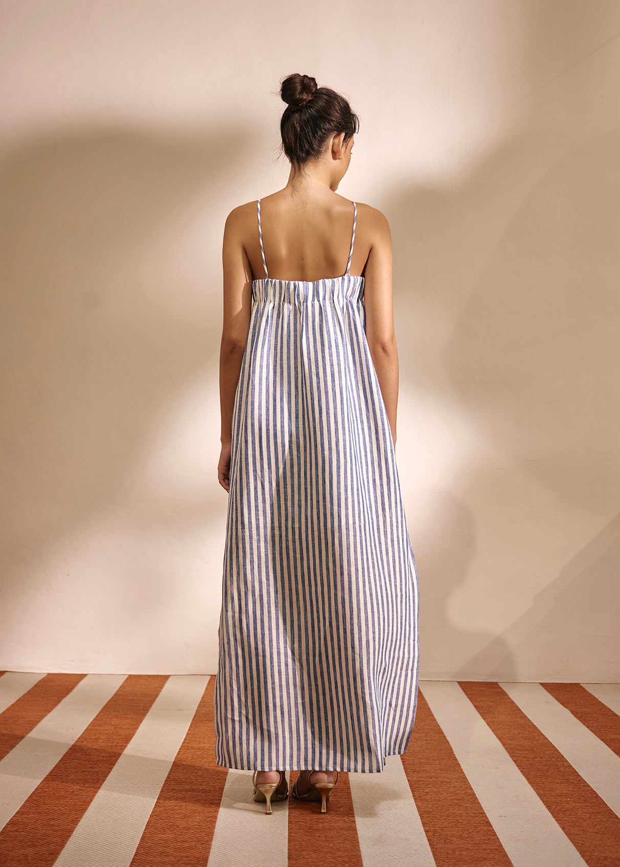 A woman viewed from behind wearing a sleeveless maxi dress with thin vertical blue and white stripes, standing on a striped floor against a plain beige wall.