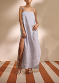 A woman modeling a sleeveless maxi dress with vertical blue and white stripes, tie straps, and a thigh-high slit, standing in a studio with a beige backdrop and striped floor.