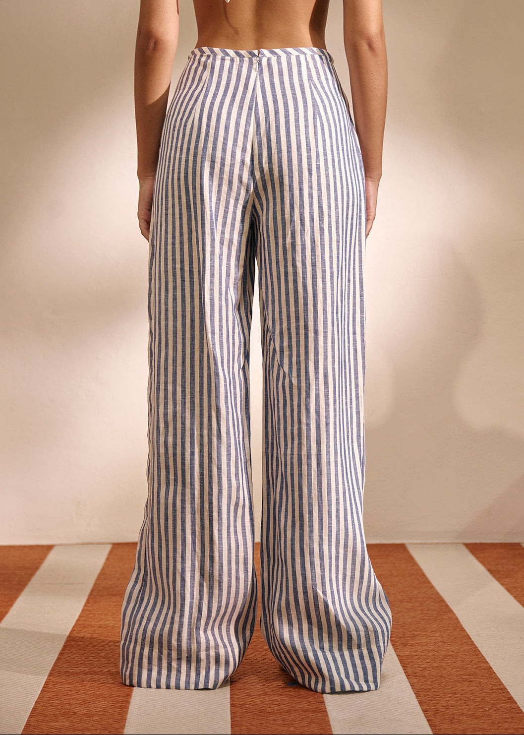 A model shown from the back wearing high-waisted, wide-leg trousers with vertical navy and beige stripes, paired with a white tie-strap halter top against a neutral studio backdrop.
