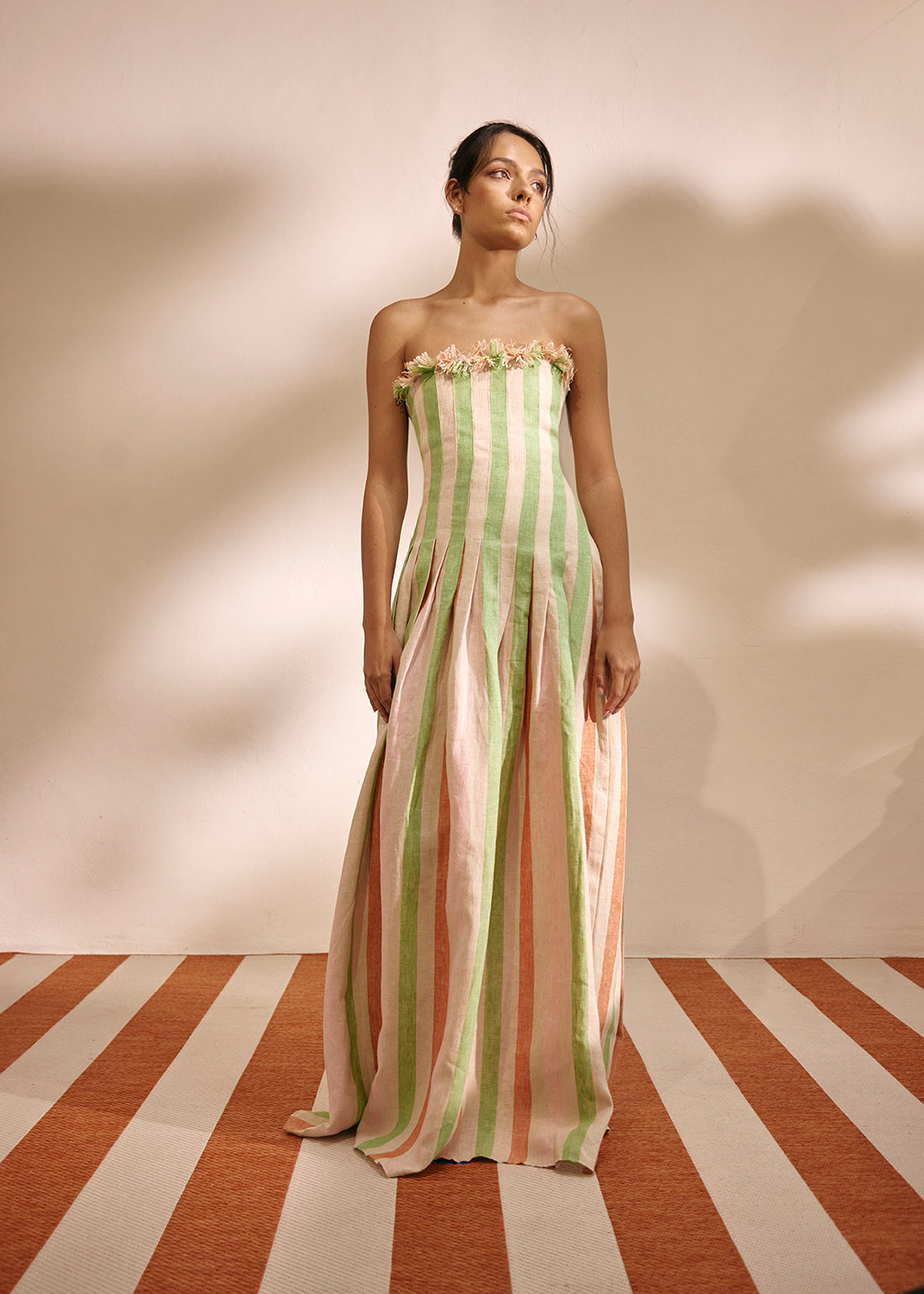 A woman stands in a studio wearing a strapless maxi dress with vertical pastel stripes in light green, peach, and cream, with a ruffled floral neckline. The background is a warm beige wall with soft shadows, and the floor has alternating cream and terracotta diagonal stripes.