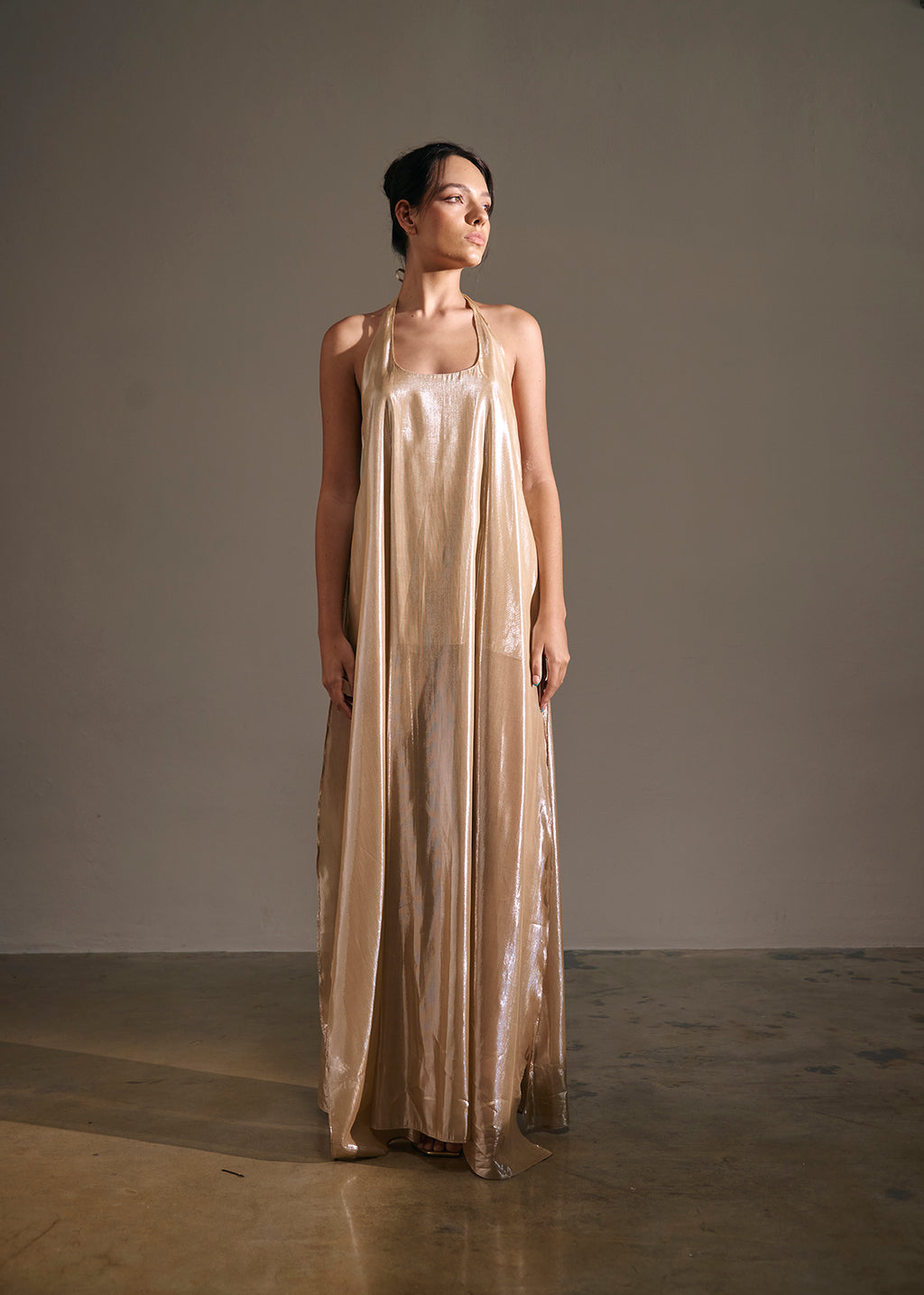 A model stands in a studio wearing a floor-length, sleeveless metallic champagne gown with a draped, flowing silhouette.