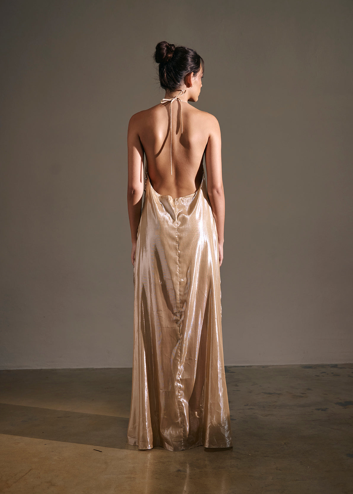 A woman stands with her back to the camera wearing a floor-length metallic champagne gown with an open back, photographed in a minimalist studio setting.