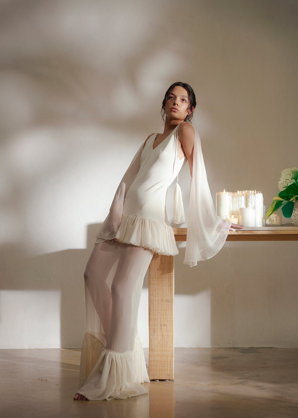 Woman in a white dress standing in a softly lit room with a table and plant.