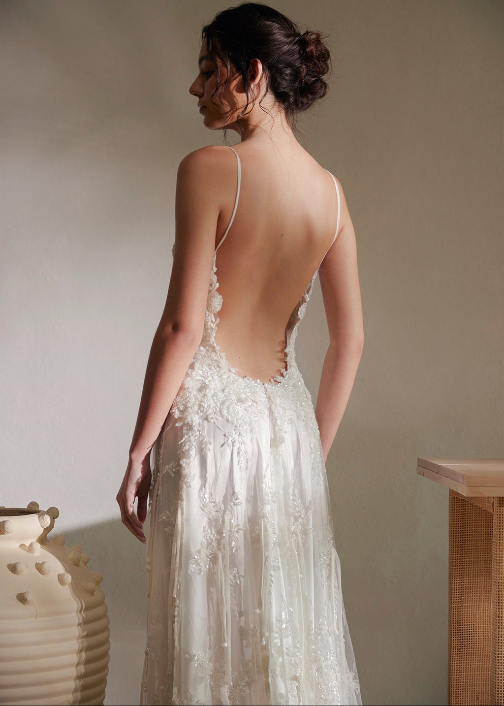 Woman wearing a white lace wedding dress with an open back, standing against a neutral wall.
