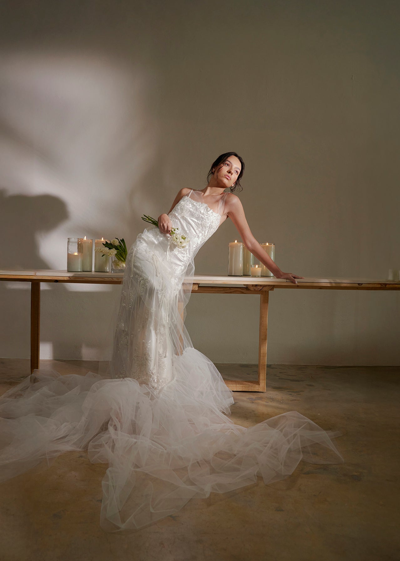 Woman in a white wedding dress standing in a minimalistic room with candles and flowers.