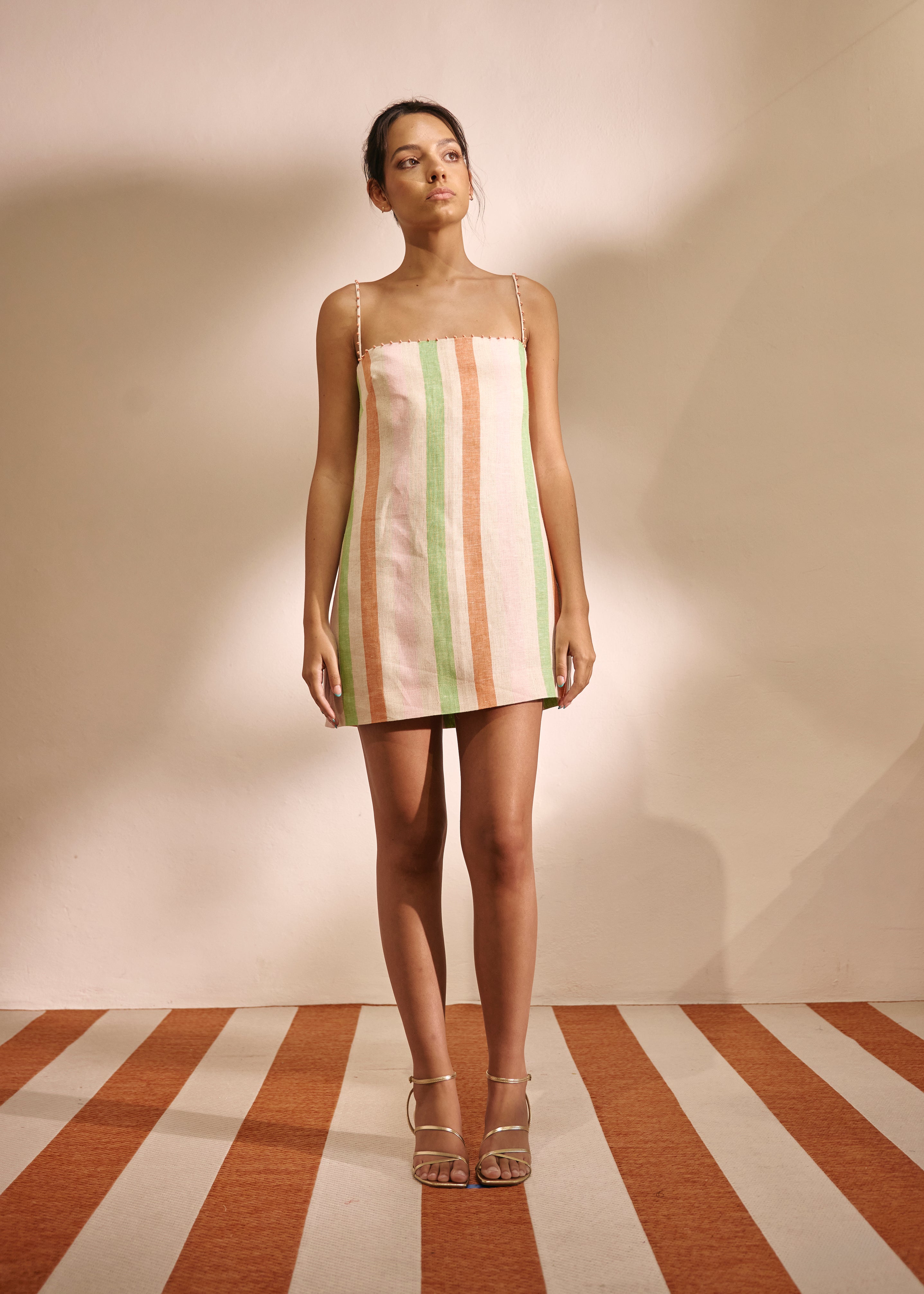 A woman stands in a studio wearing a sleeveless mini dress with vertical pastel stripes in cream, mint green, and peach. The background is a warm beige wall with strong shadows, and the floor has matching beige and rust-orange stripes.