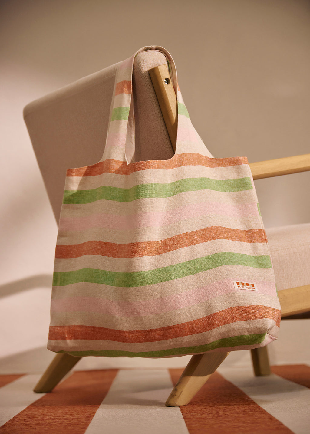 A striped canvas tote bag with horizontal bands of peach, sage green, and light pink on an off-white base, hanging from the arm of a mid-century modern wooden chair.