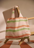 A striped canvas tote bag with horizontal bands of peach, sage green, and light pink on an off-white base, hanging from the arm of a mid-century modern wooden chair.