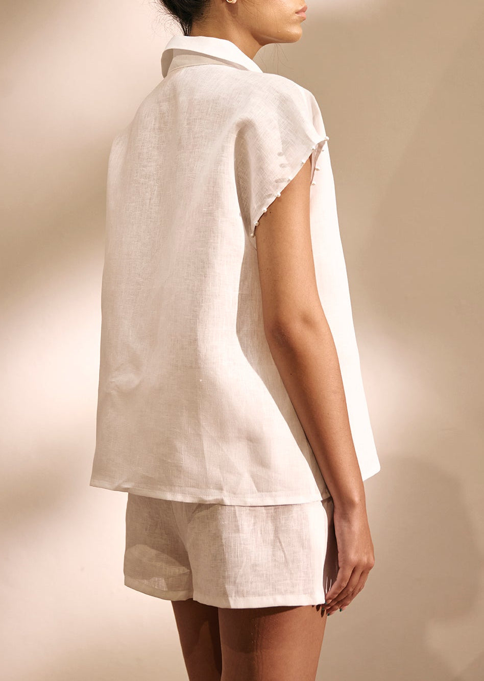 A woman shown from the back wearing a loose white linen shirt and matching shorts against a warm beige studio background.