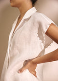 A close-up of a model wearing a white linen shirt with short sleeves, a pointed collar, and decorative buttons along the sleeve cuffs, set against a neutral beige background.
