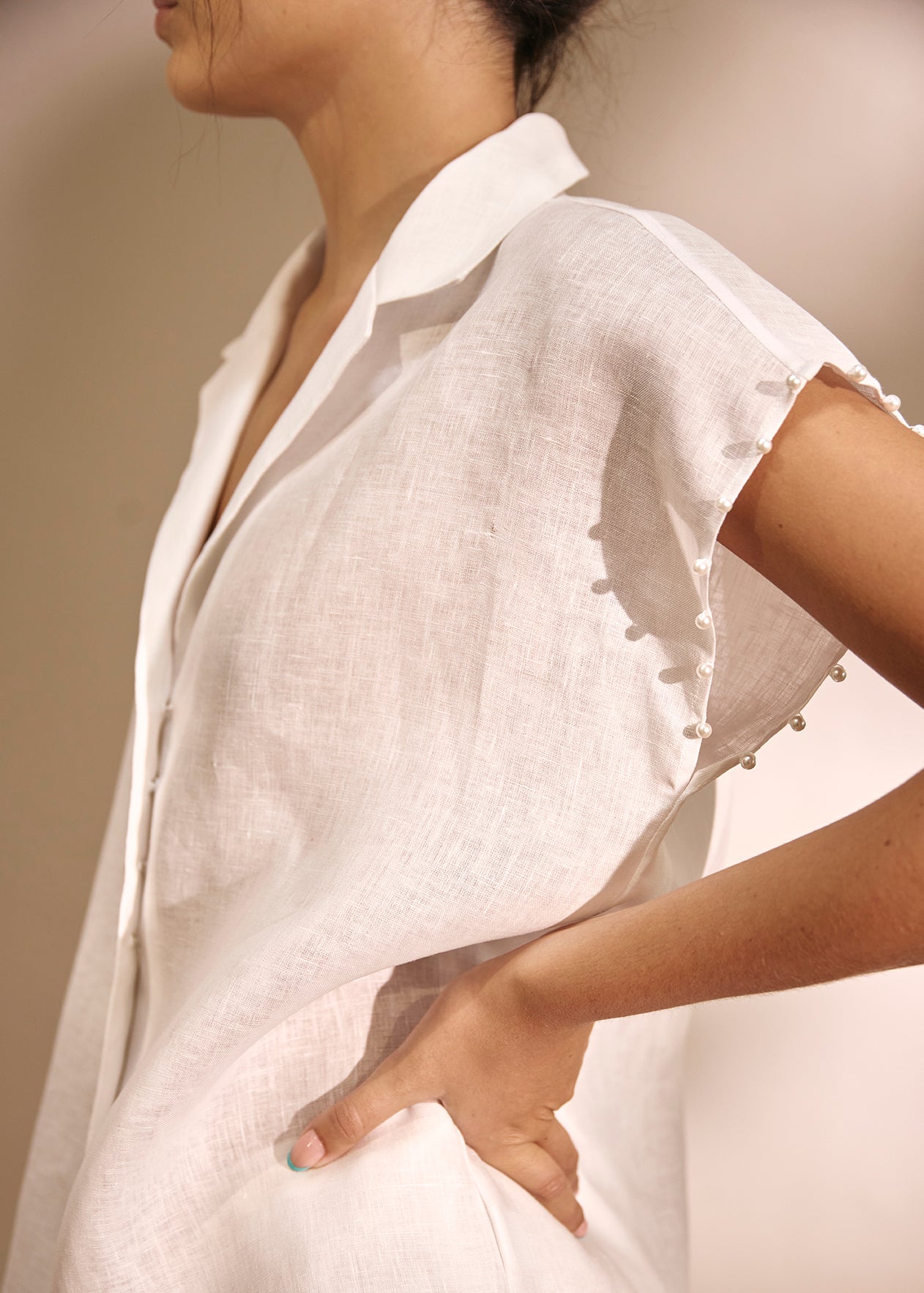 A close-up of a model wearing a white linen shirt with short sleeves, a pointed collar, and decorative buttons along the sleeve cuffs, set against a neutral beige background.