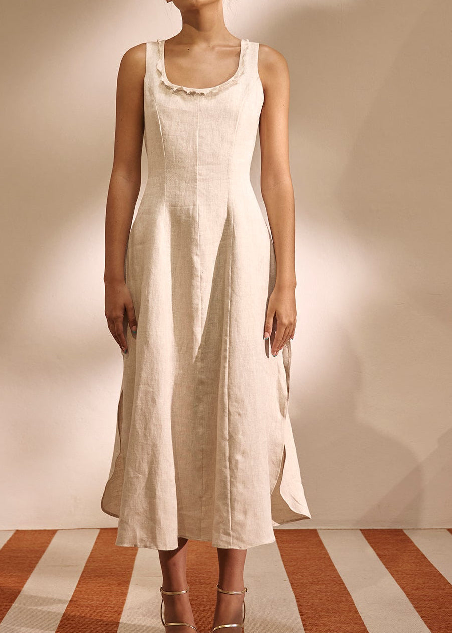 A woman modeling a sleeveless, off-white linen midi dress with a ruffled neckline, standing on a striped terracotta and cream floor against a warm beige wall.