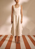 A woman stands in a studio wearing a sleeveless, midi-length cream linen dress with a ruffled neckline. The background is a warm beige wall with soft shadows, and the floor features alternating vertical stripes of terracotta and cream.