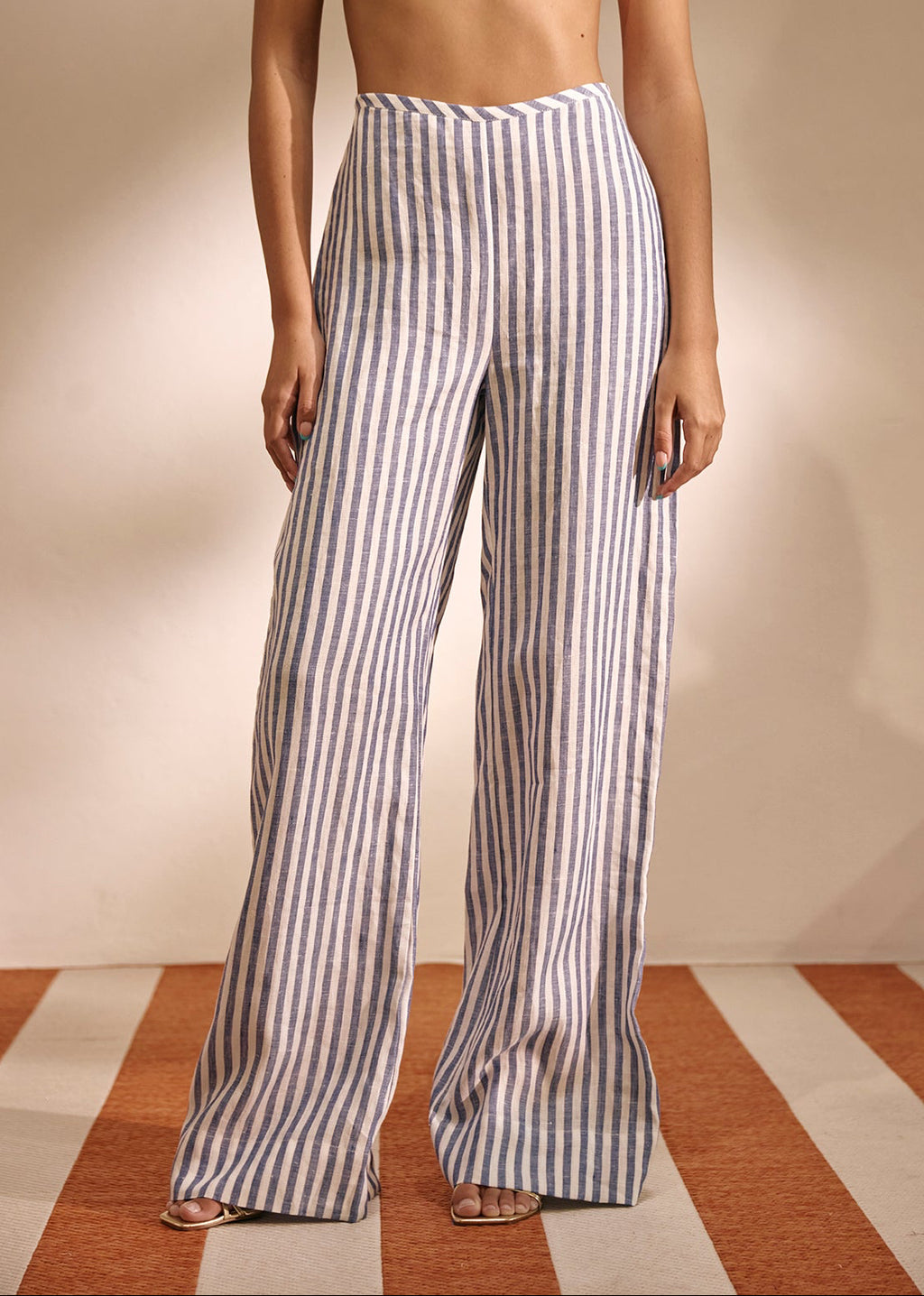 A model wearing high-waisted, wide-leg trousers with vertical blue and white stripes, paired with a cream bikini top, standing against a beige wall on a striped floor.